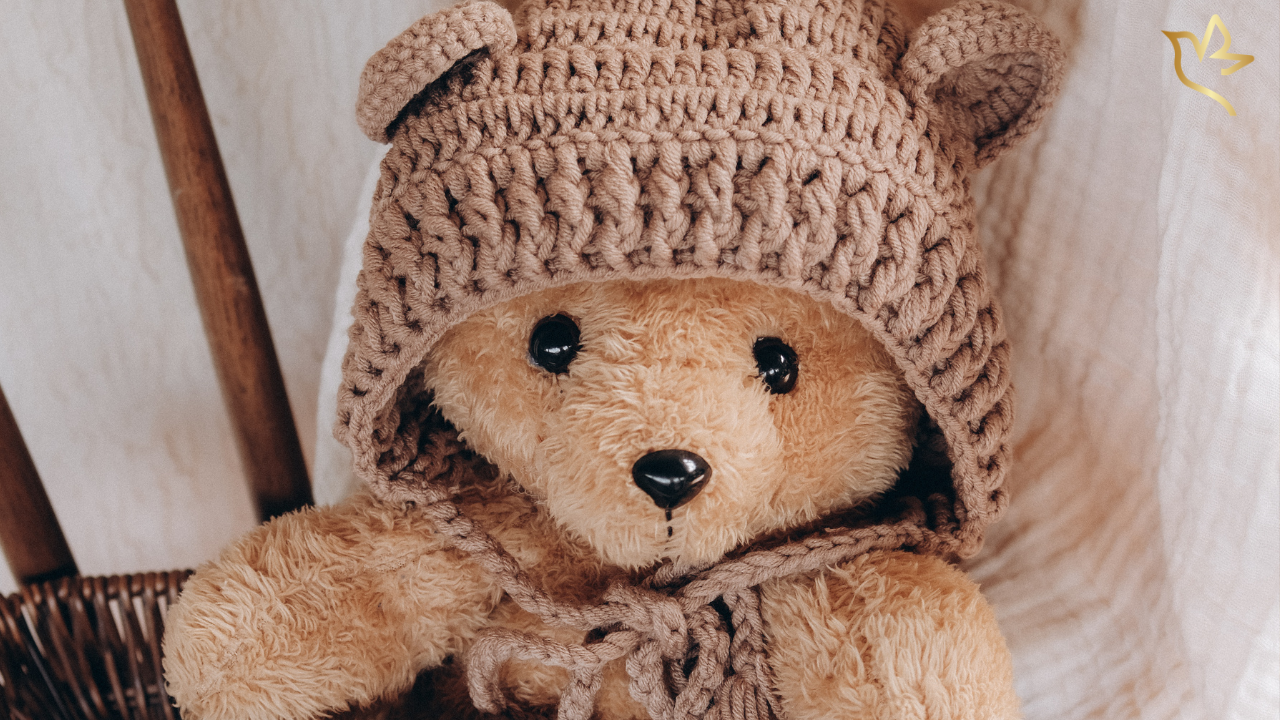 A well-loved teddy bear wearing a knitted hood rests quietly in a wicker chair, symbolizing the comfort, memories, and enduring love found in treasured keepsakes.