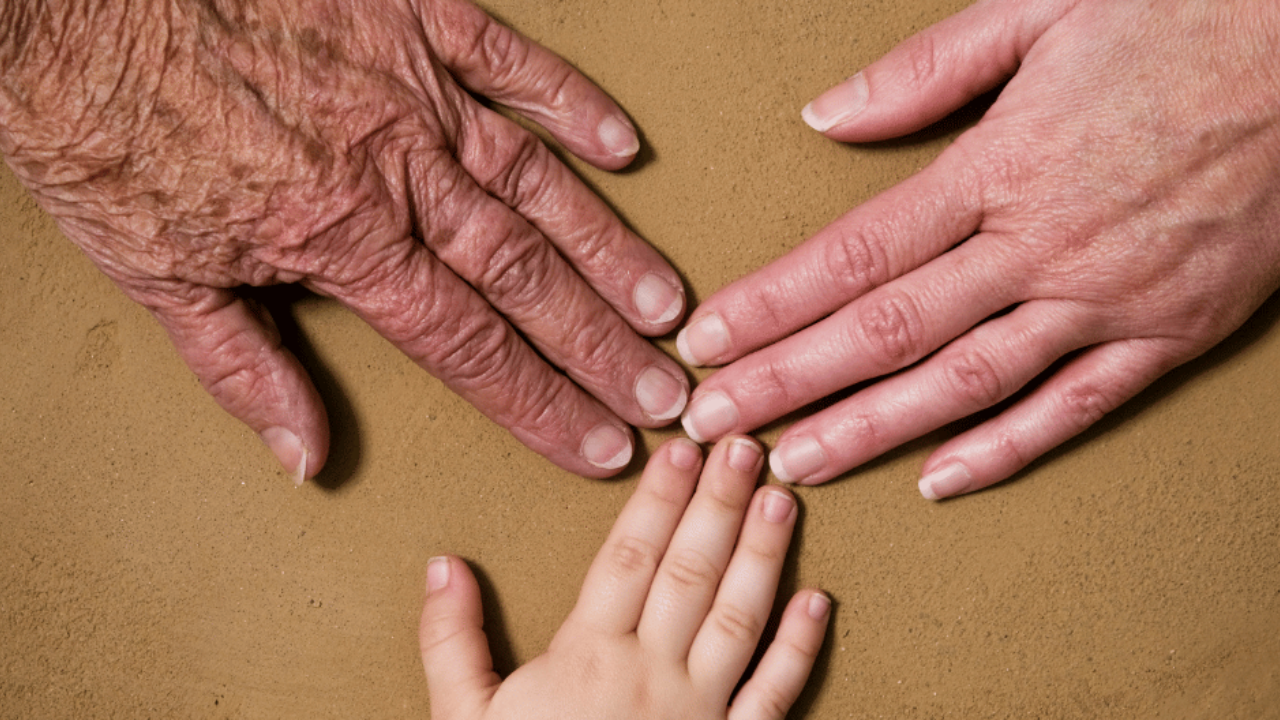 Elder, adult, and child hands touching fingertips in sand, representing three generations and generational trauma healing
