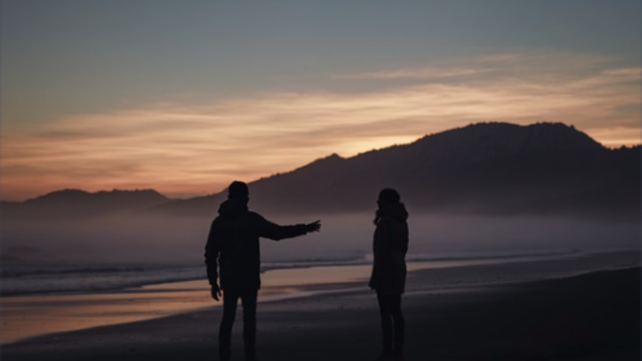 Two silhouetted figures on a foggy beach at dusk, one reaching toward the other, symbolizing emotional distance in relationships