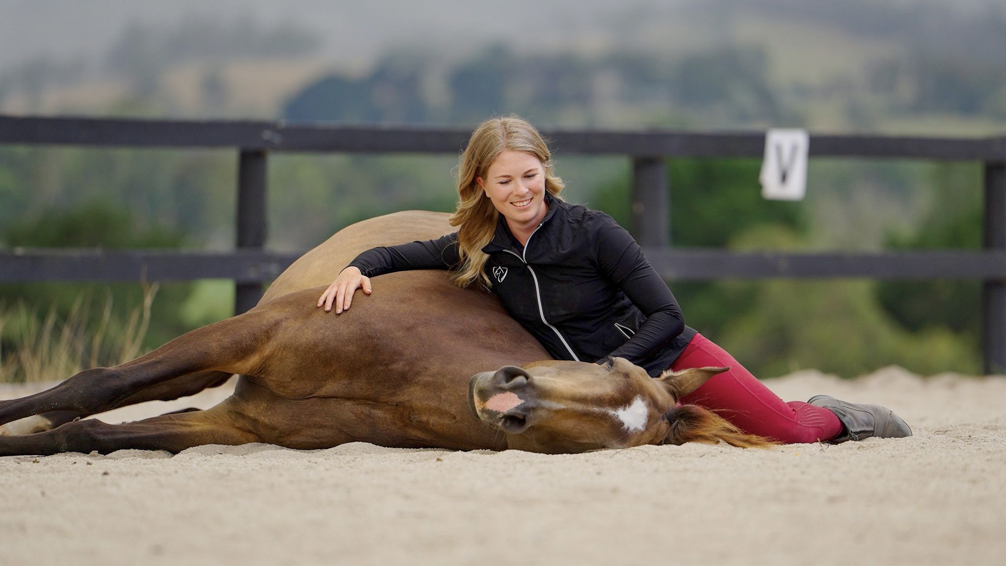Nadine lying in the sand arena, resting against her chestnut horse as he lies down beside her