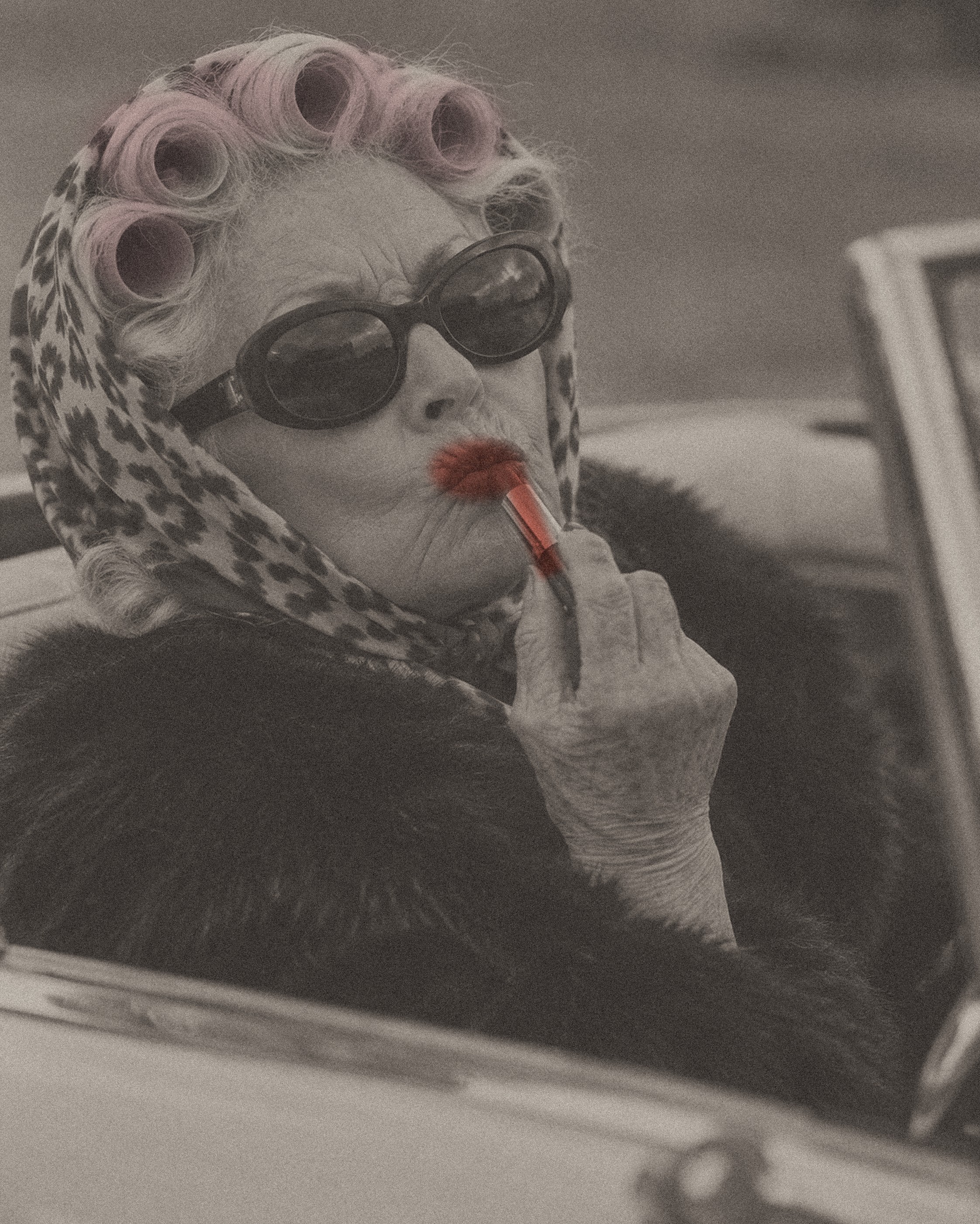 An older woman in sunglasses and hair rollers, applying lipstick