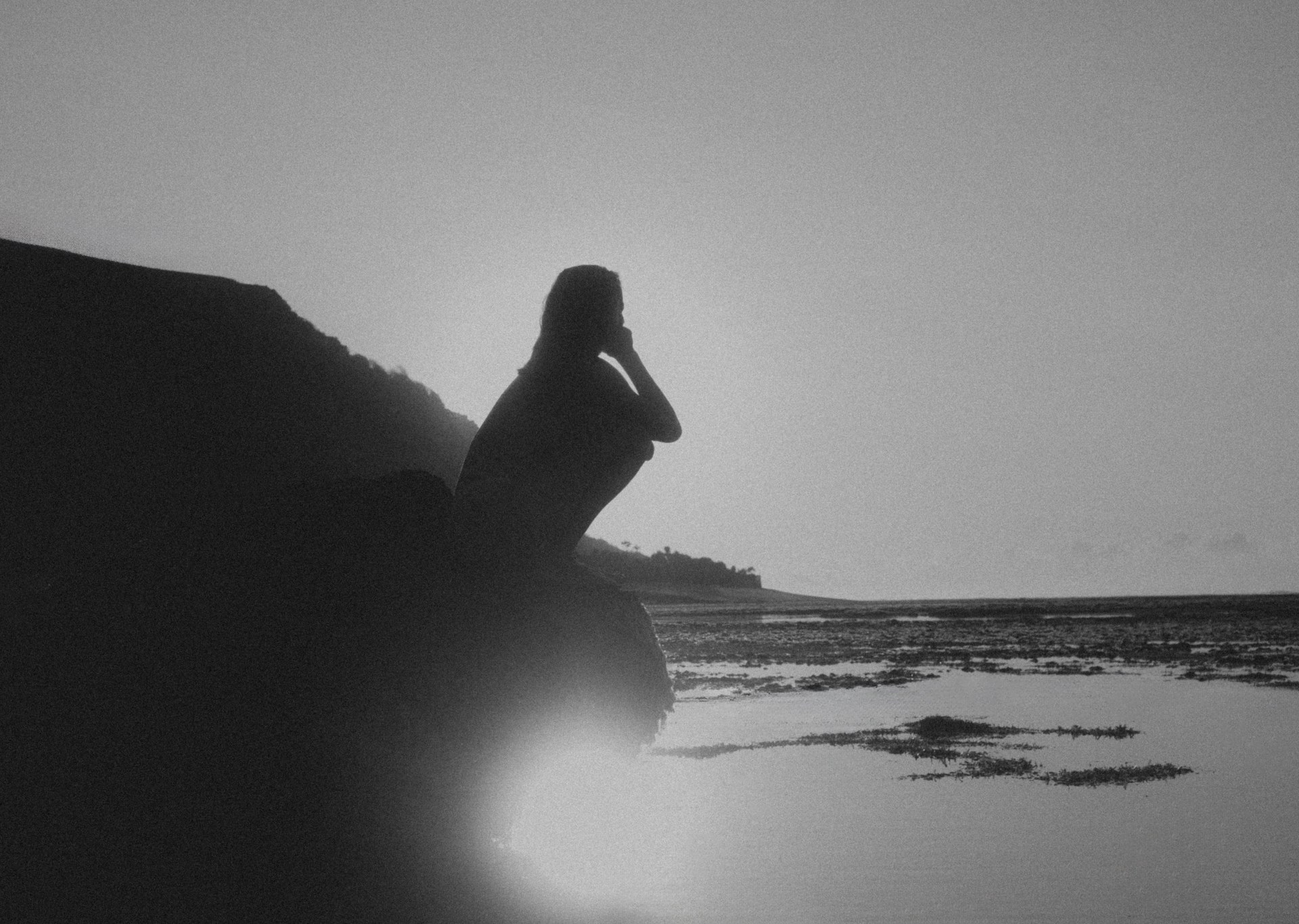 Rachel in silhouette, crouched on a rock by the sea, hand to her face