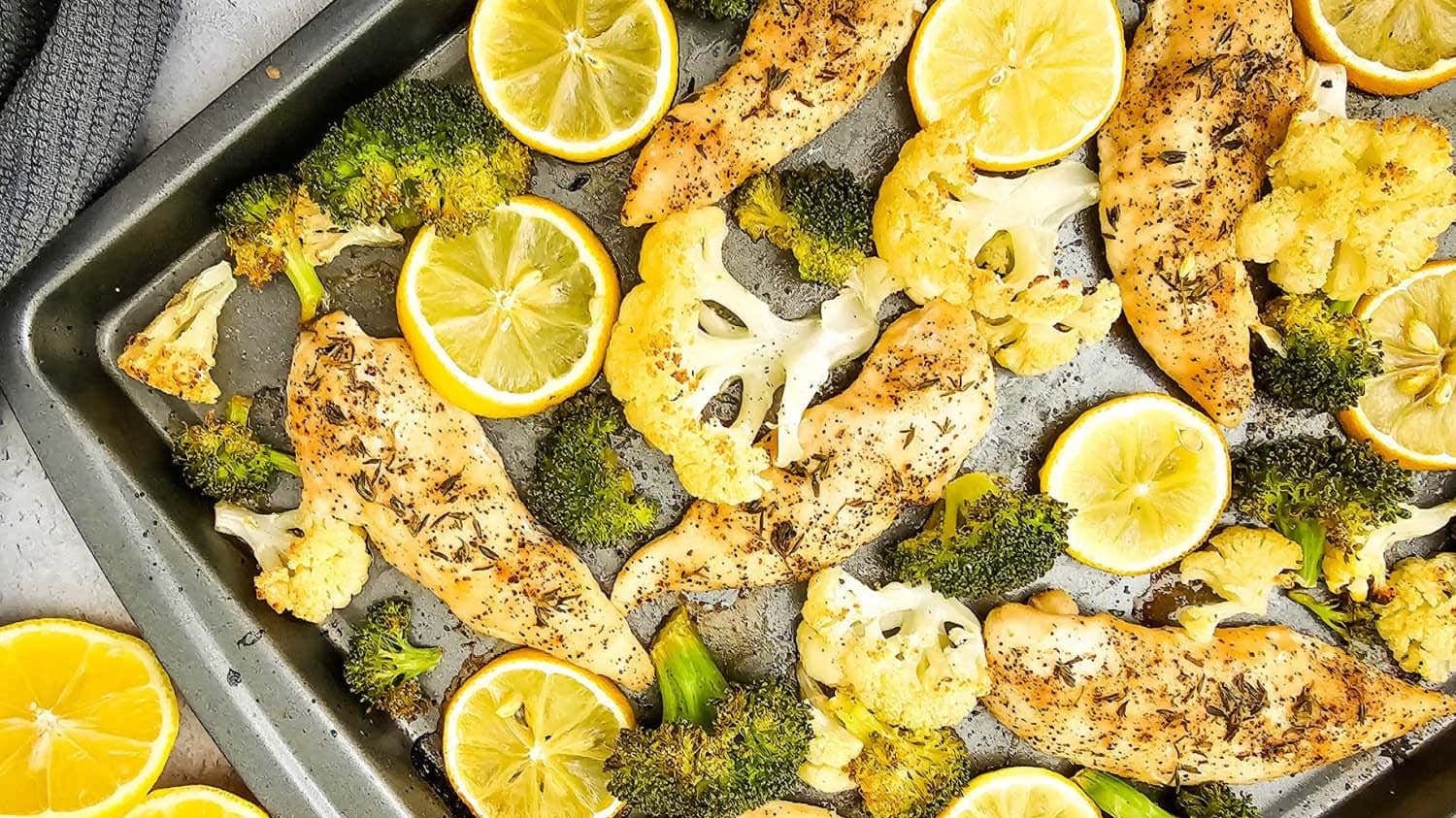 Roasted cubed chicken breast and green broccoli florets arranged on a parchment-lined baking sheet pan with lemon slices.