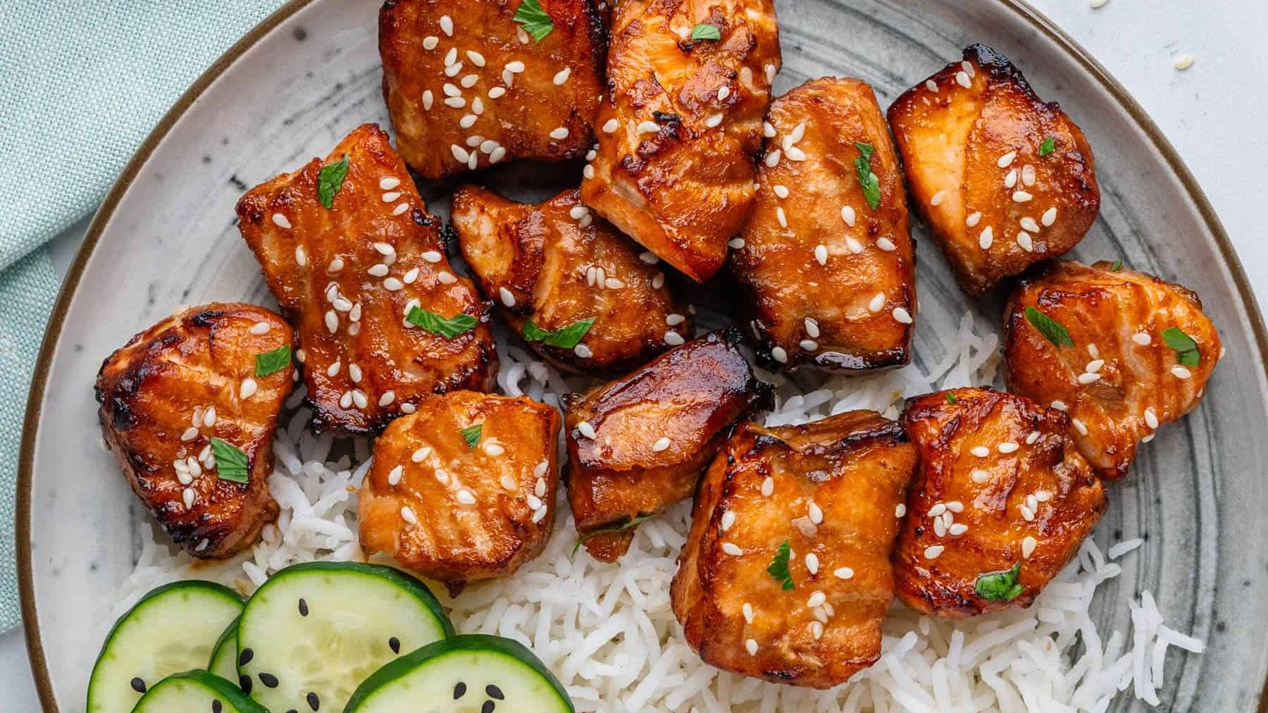 Seared cubes of salmon glazed in a glossy brown sweet chili sauce inside a non-stick skillet, garnished with white sesame seeds and green onions.