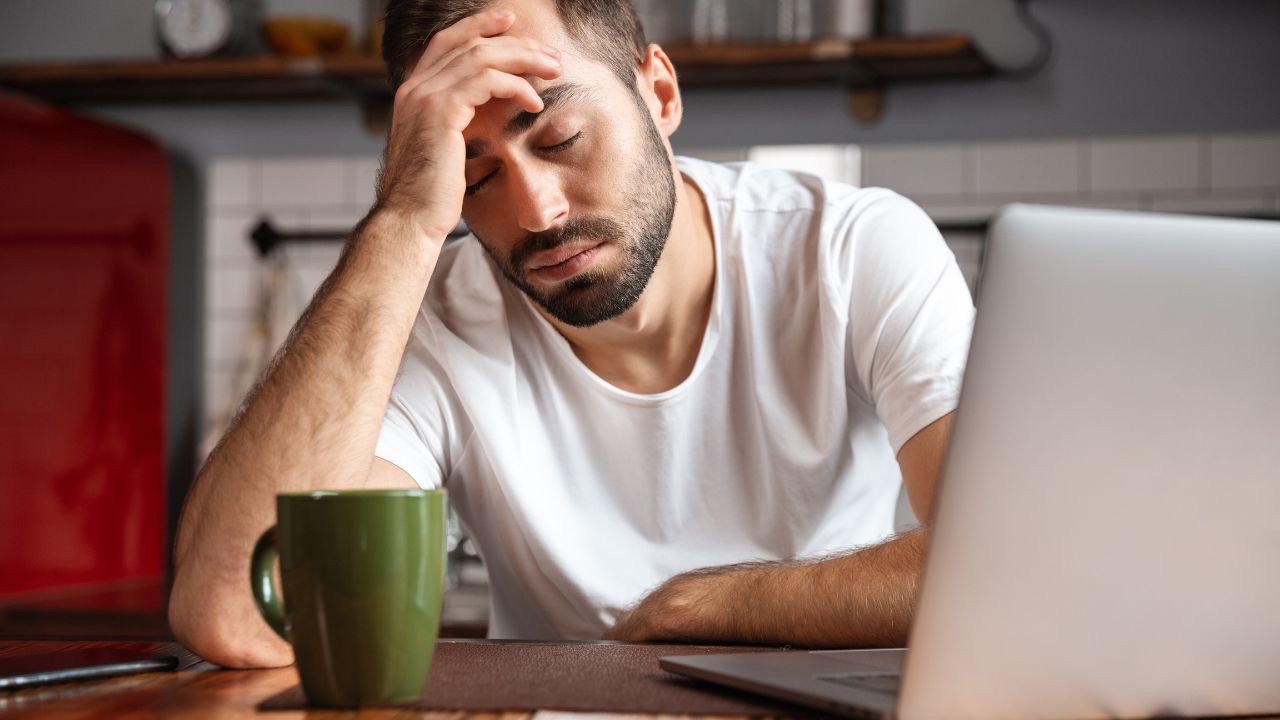 Image is of a person wearing a white short sleeve tee shirt sitting at table in front of a laptop. There is a green coffee mug on in front of them, and they have their eyes closed and on hand on their head. Google is good for many things, but it can't tell you what it's like to be the parent of a transgender child. 