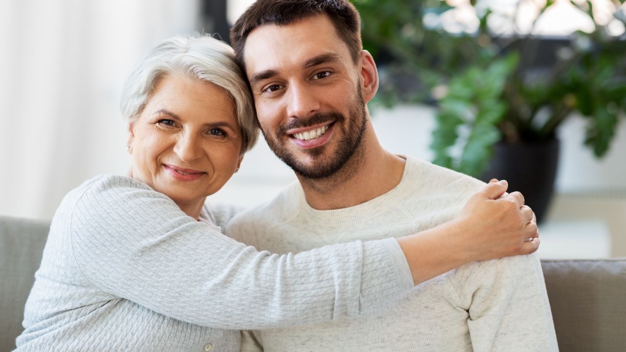 Image is of a two people sitting on a couch. The parent is wearing a light blue long sleeve shirt, and has short, white hair. Their arms are wrapped around their adult child, who has dark hair, a beard and mustache and is wearing a white long sleeved tee shirt. Both people are smiling. 