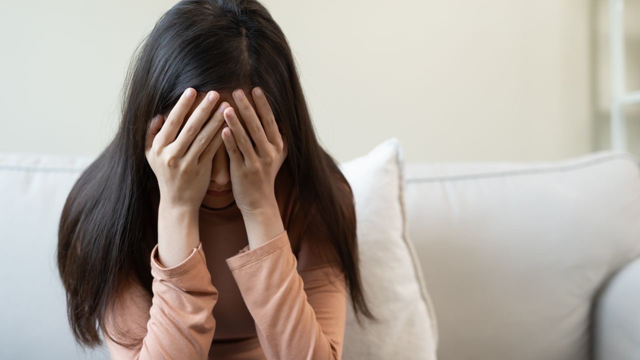 Image is of a person with long dark hair and a long sleeve peach teeshirt sitting on a white couch with a white pillow behind them with their arms up and both of their hands covering their face. It's difficult navigating complicated relationships when your child comes out as transgender. 