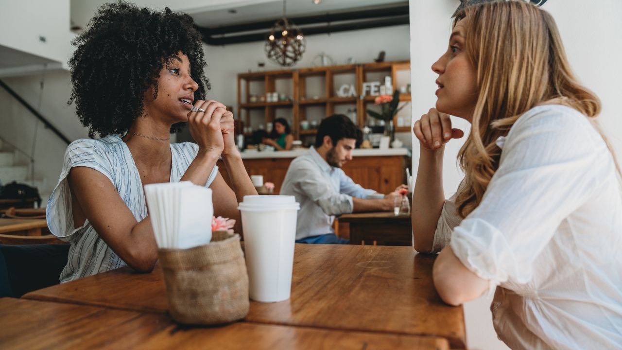 Image is two people sitting in a coffee shop facing each other. Both have their hands up in front of them, and they are deep in conversation, engaged in listening. 