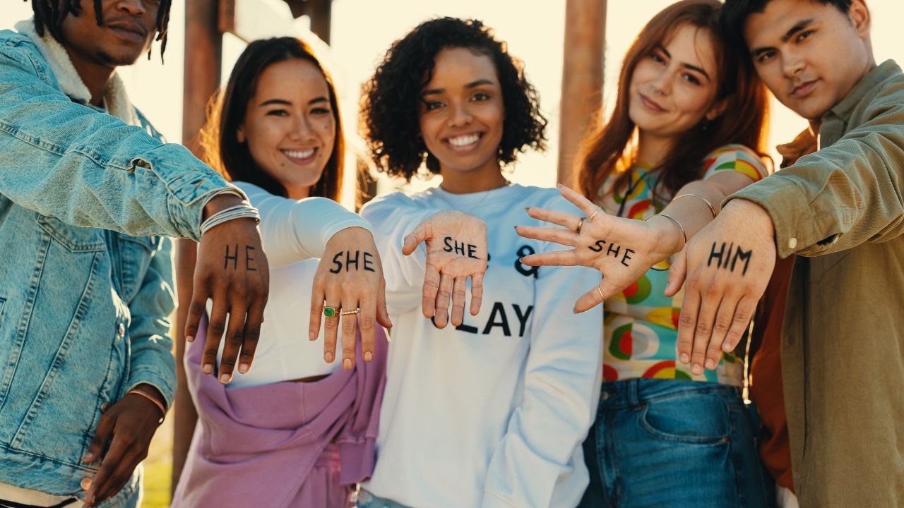 Image is of several people standing outside with their hands all facing forward, and a pronoun written on each hand. In order from left to right, they read He, she, she, she, him. Each person is smiling. 