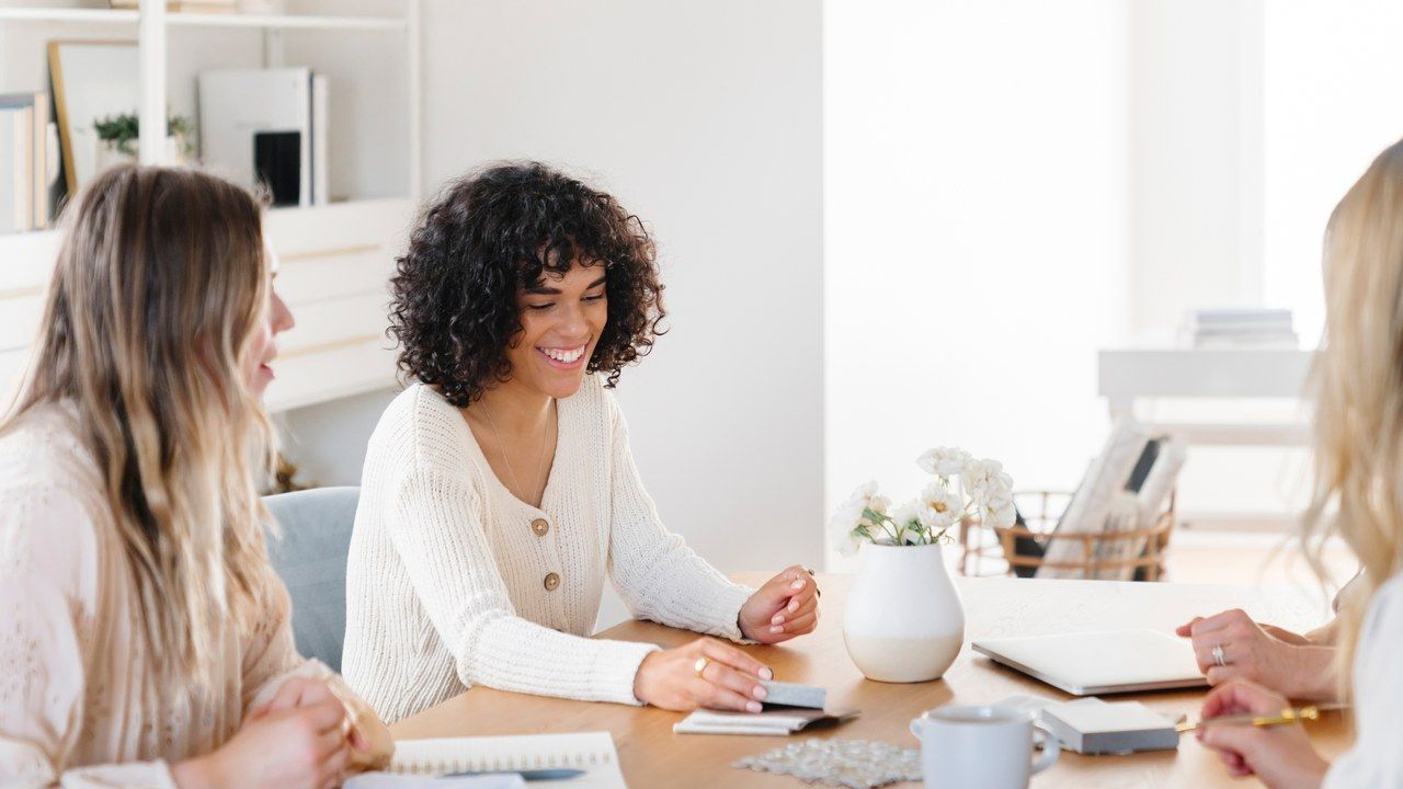 Three women sitting around a wood table with notebooks and coffee, mid-conversation.