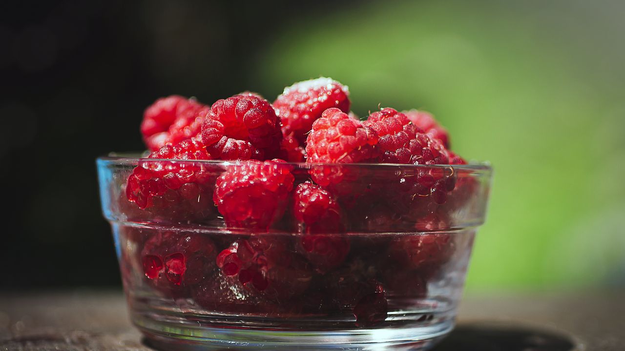 Glass bowl piled high with fresh ripe raspberries sitting on a wooden table in warm sunlight