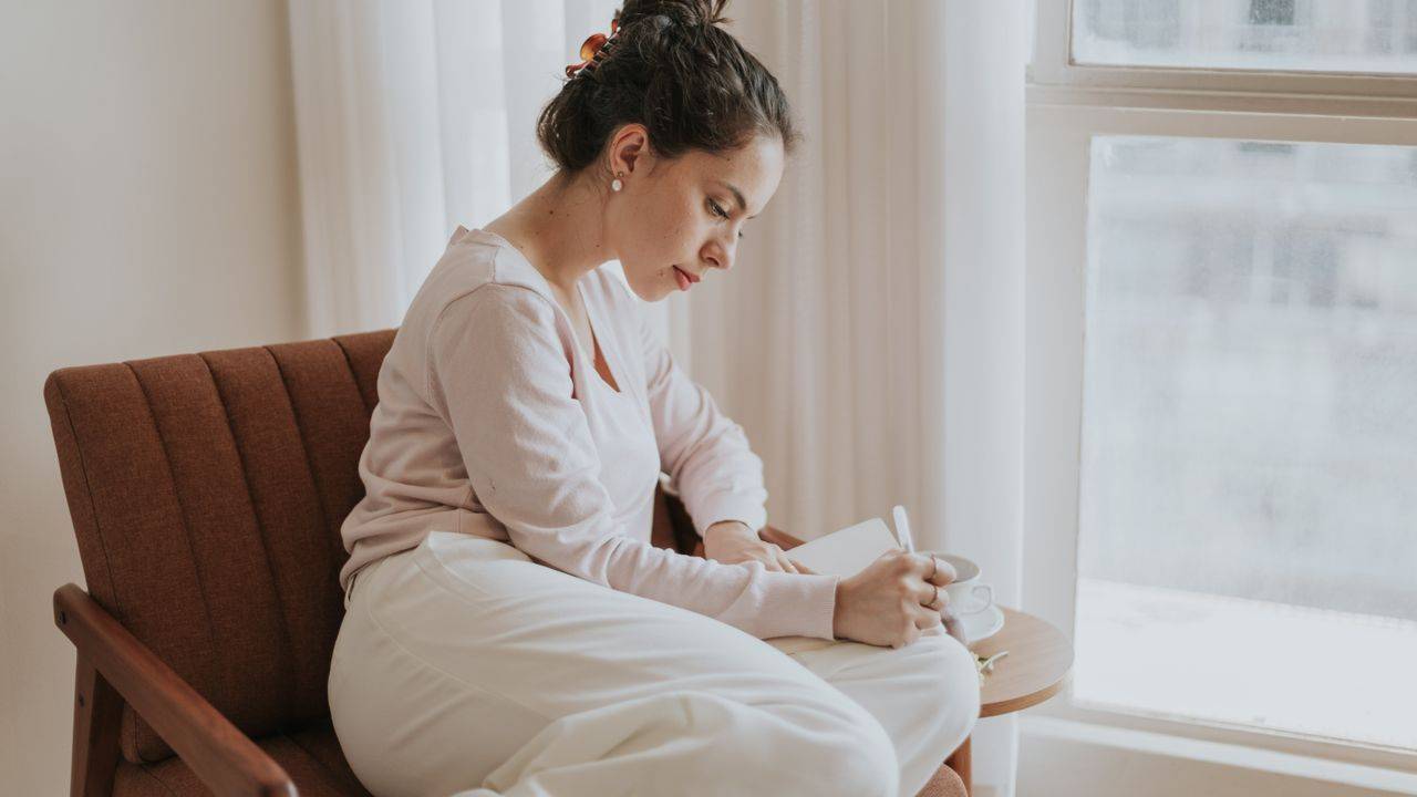 Woman in a cream jumper sitting in a brown armchair writing in a notebook by a bright window