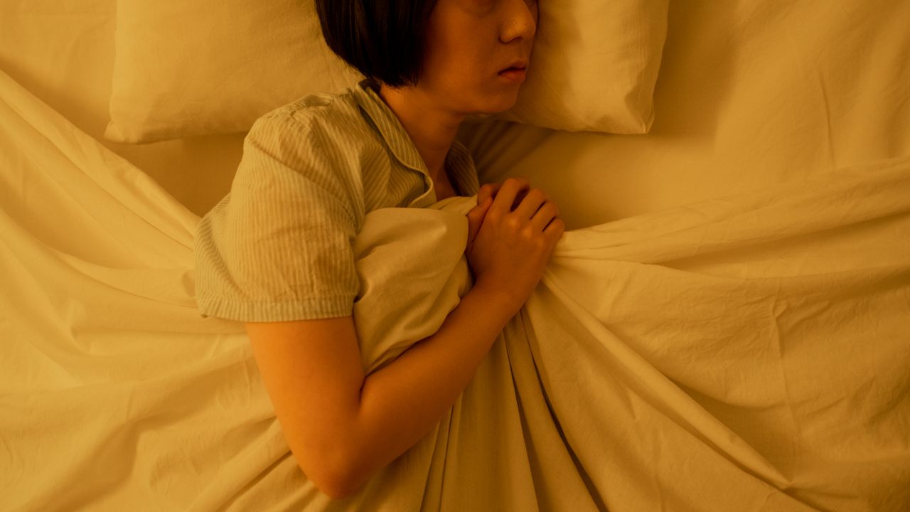 Woman lying awake in bed under warm lamplight, clutching the sheets as she struggles to sleep at night