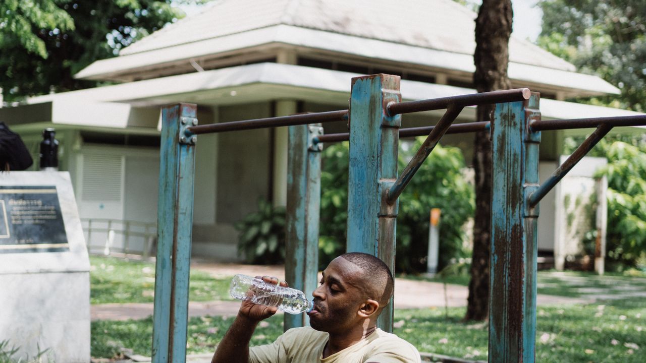 Man in a beige T shirt sitting by outdoor pull up bars in a leafy park, drinking from a clear bottle of water after a workout