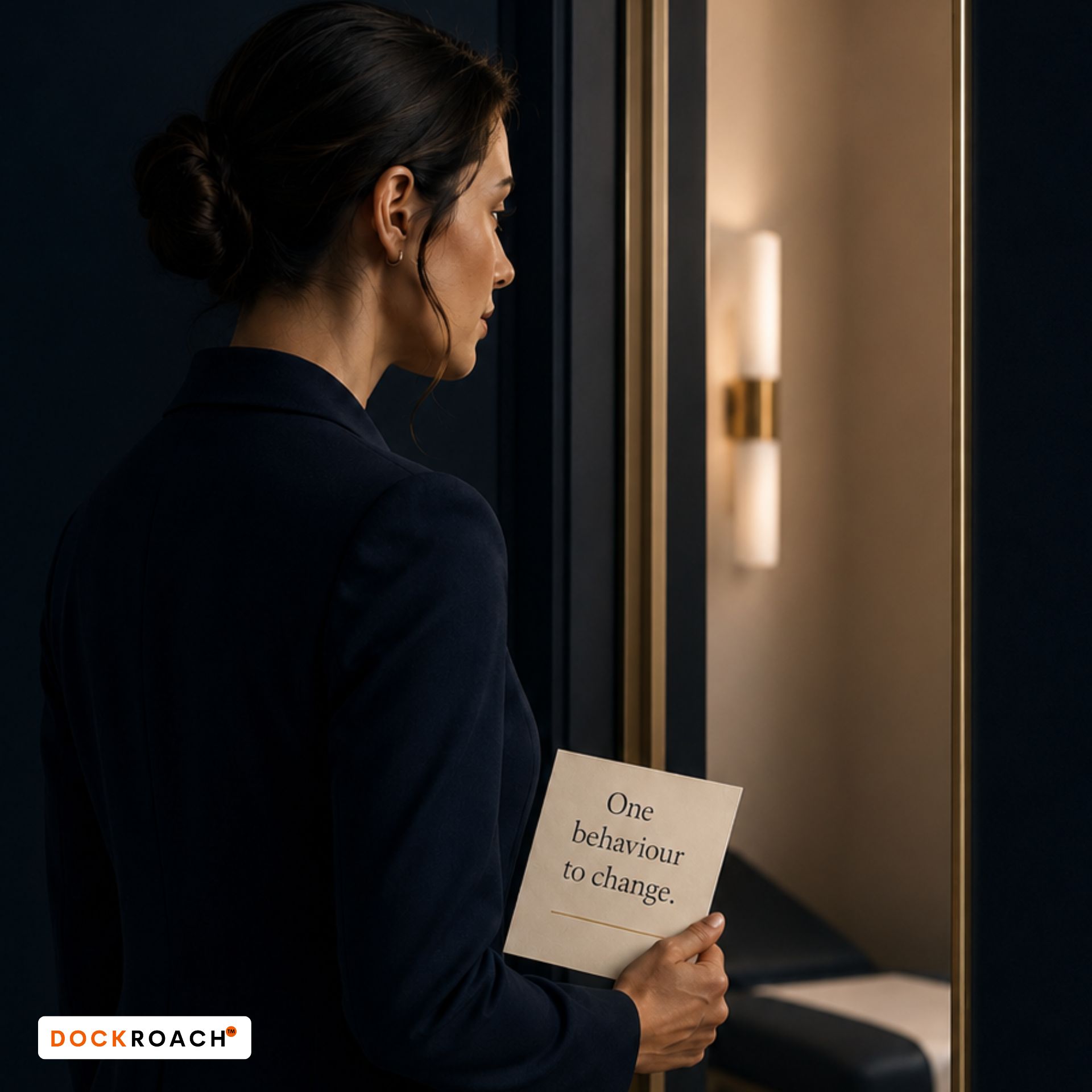 One Behaviour to Change A medical candidate stands outside a clinical station doorway holding a card that reads “One behaviour to change,” representing feedback being turned into focused action.
