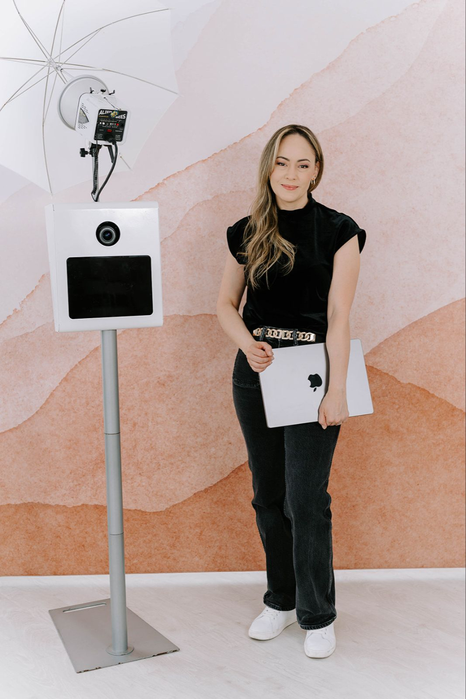 a woman is stilling on a stool facing to the right. She is smiling and her hair is falling infront of her face. She has her left arm up behind her head. In the background is a beige hand painted backdrop. 