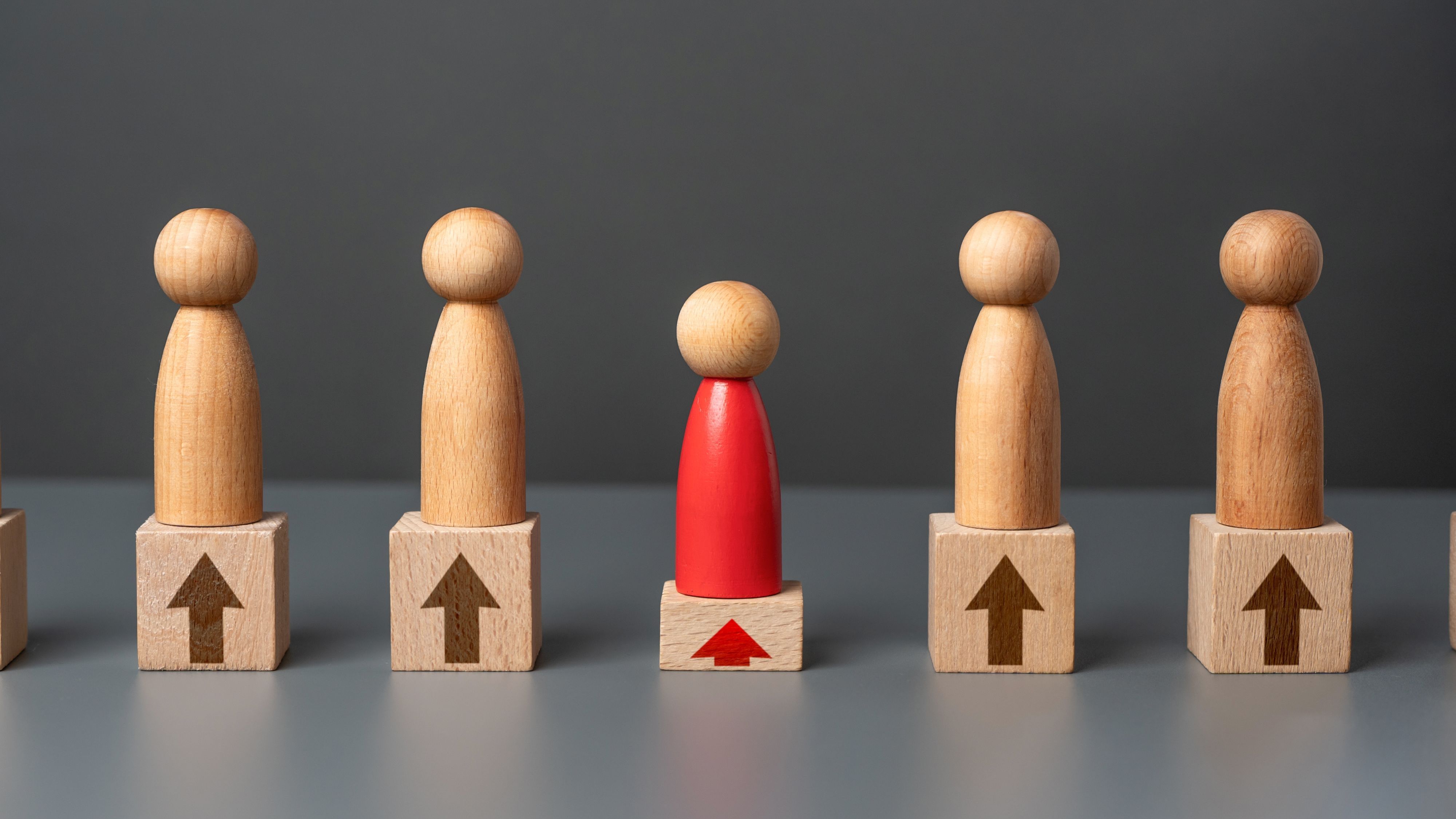 A red wooden figure standing among plain wooden figures on blocks with upward arrows, symbolising the impact of a strong Integrator versus “good enough” performance in EOS‑run businesses.