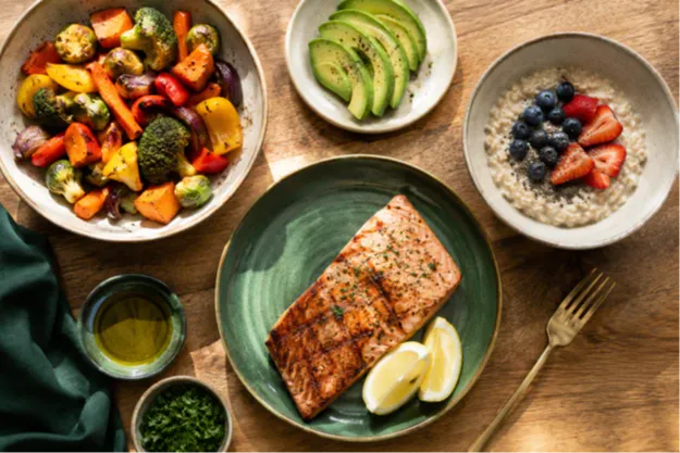 An overhead photo of a balanced, real-food meal spread including salmon, roasted vegetables, avocado, oats, and olive oil on a warm wooden table. This is what simple eating for weight loss looks like in real life: colorful, nourishing, and completely doable.
