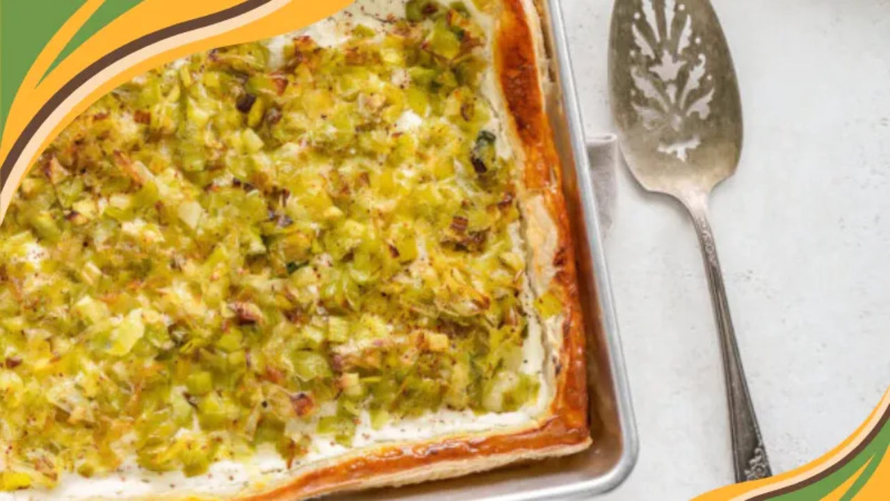 Caramelized leek and ricotta tart baked in a rectangular sheet pan, topped with golden-brown leeks and a creamy ricotta filling. The tart has a crisp, flaky pastry crust with lightly browned edges and is photographed from above beside a vintage serving spatula on a light countertop.