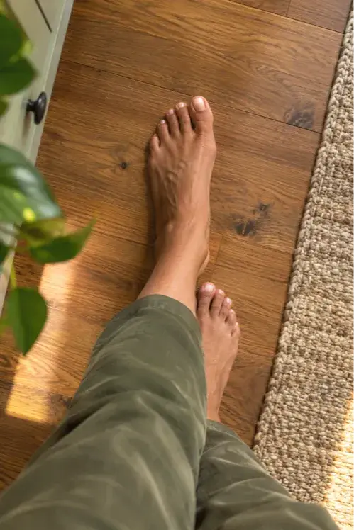 A close view from above of bare feet placed heel to toe on a warm wooden floor. The pose is the heel to toe walk, the second balance drill in Coach Deb's four week routine.
