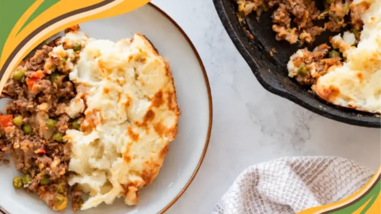 Plate of creamy shepherd's pie with mashed potatoes, next to a cast iron skillet, on a white marble surface.