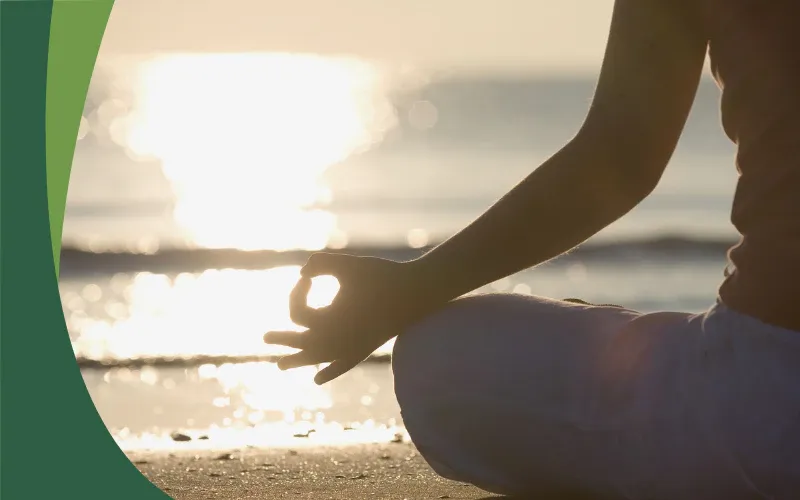 Person sitting cross-legged on a beach at sunset with one hand resting in a meditation pose. Sunlight reflects across the water, creating a peaceful and calming atmosphere.