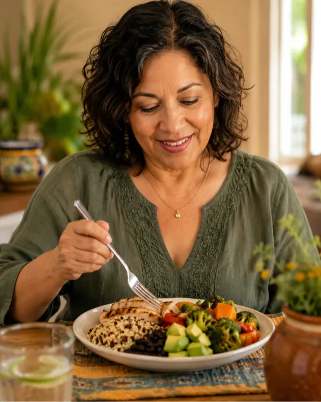A Latina woman in her late 50s enjoys a balanced, protein-rich meal at a bright dining table. Her relaxed expression reflects the ease of eating real, nourishing food. This image connects to the Get Real Weight Loss philosophy of sustainable, compassionate eating for women over 50.