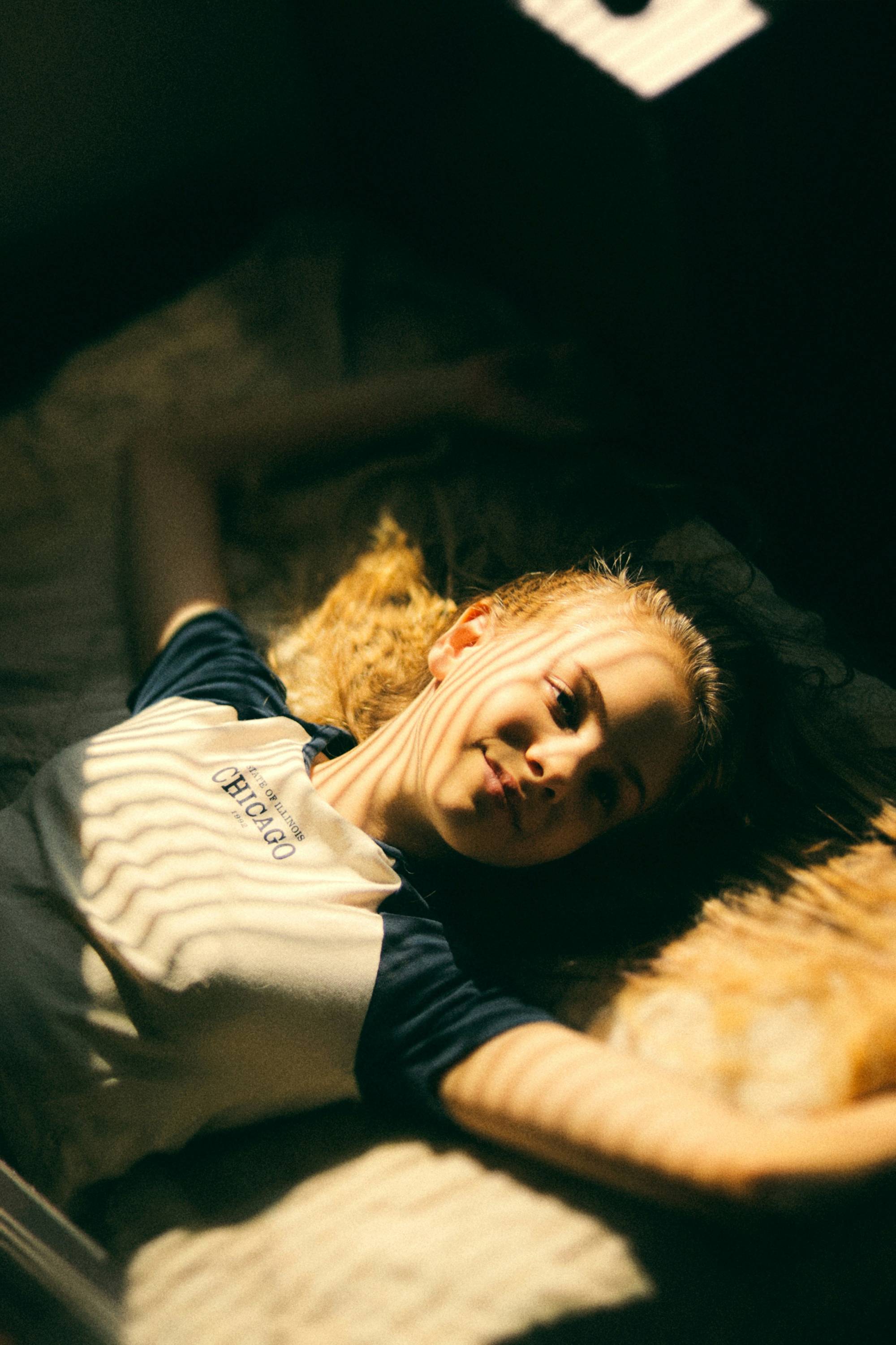 A teenager lying awake on her bed in the morning, with light from the blind falling in stripes across her face.