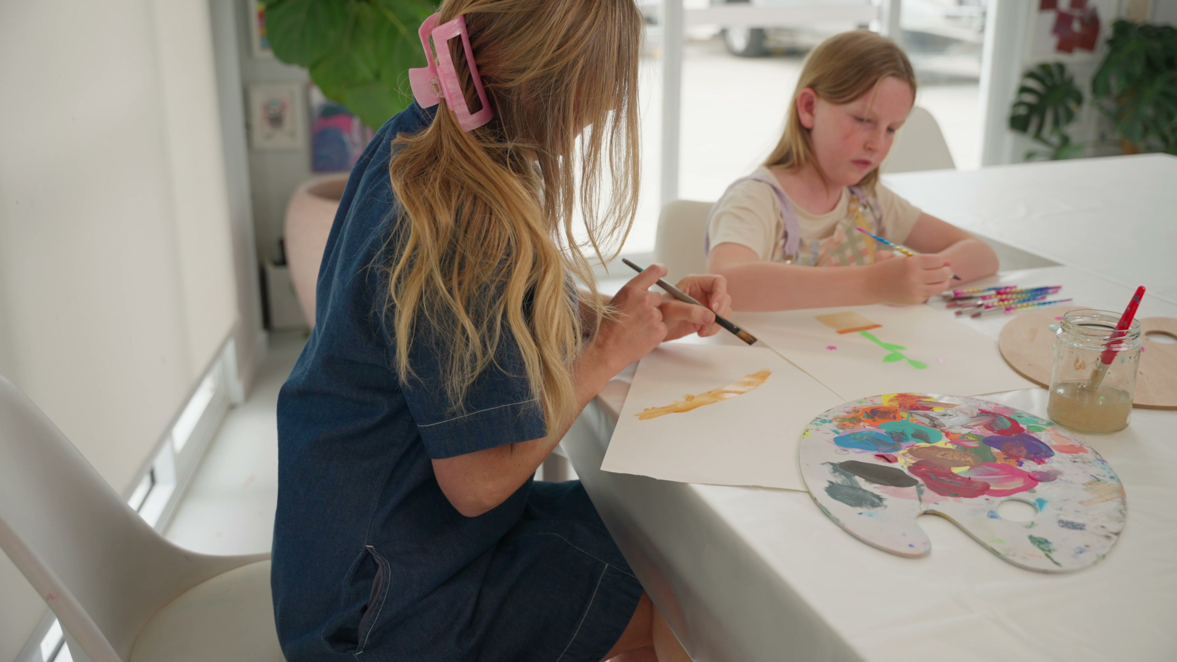 An adult sits alongside a child at a table, both painting, in a calm sunlit room
