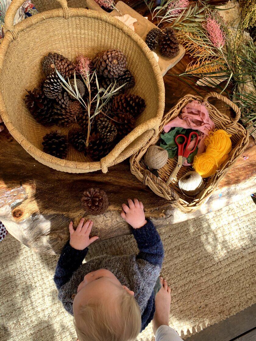 A young child reaching into a basket of pine cones on a rug