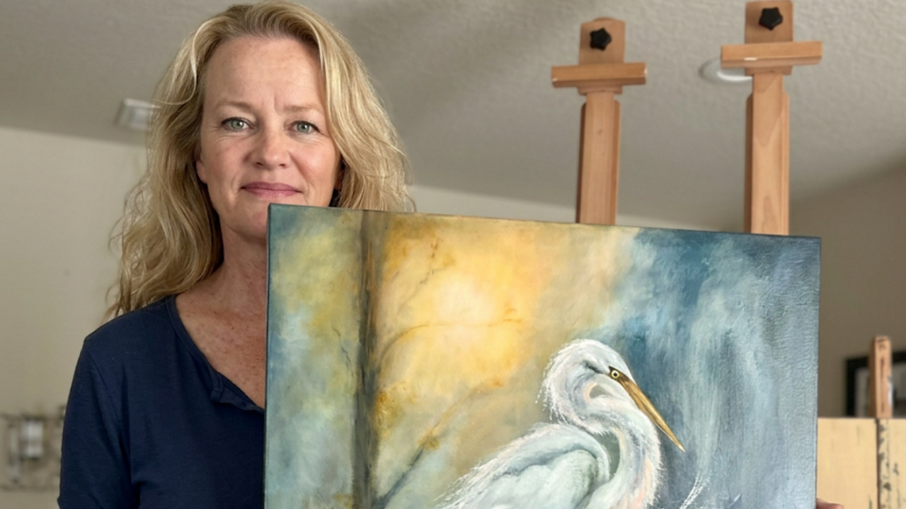 Jenn Royster holding her original oil and cold wax painting, Whispers Along the Water's Edge, featuring two egrets.