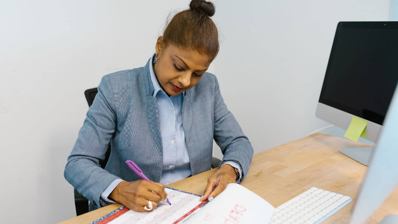 Woman in a blue blazer marks dates on a desk calendar beside a computer in an office.