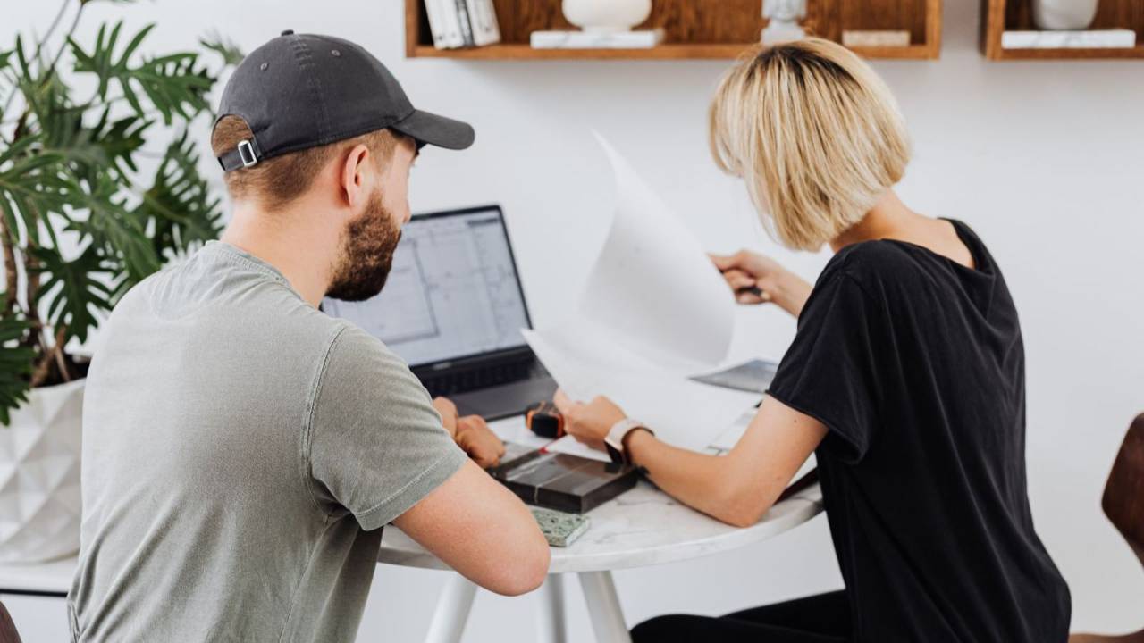 Two colleagues reviewing campaign layouts on a laptop at a shared office desk, with printed boards and notes spread across the table.