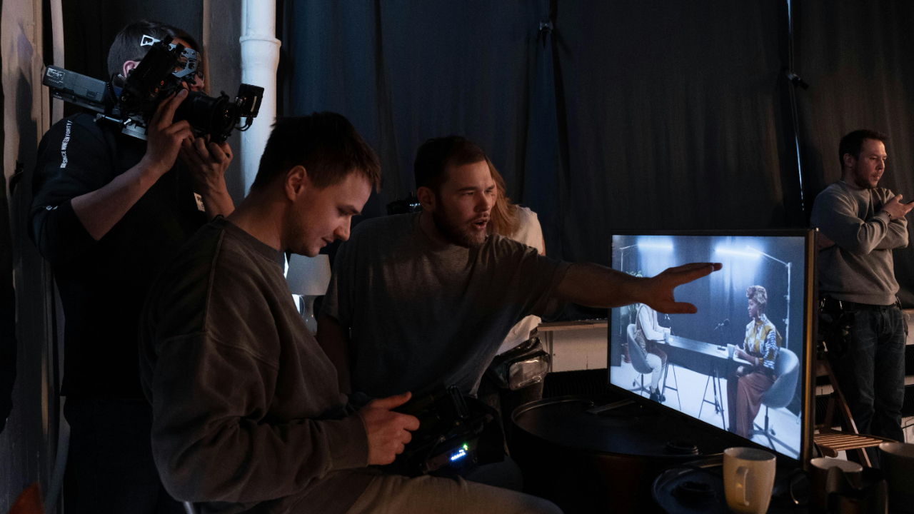 Film production team reviewing call sheets and schedules with a producer at a folding table on a working film set with camera and lighting equipment in the background