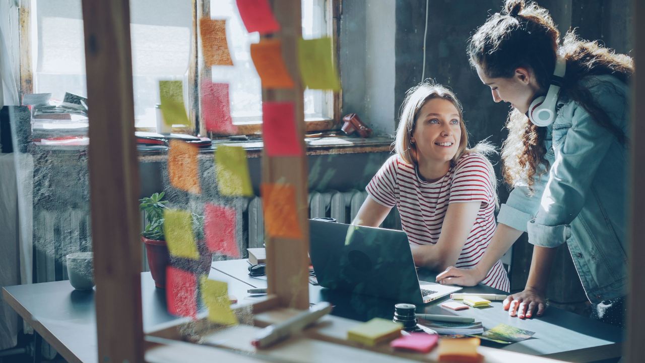 Creative project manager and producer collaborating over a production schedule and notes at a shared desk with laptops in a bright modern agency office
