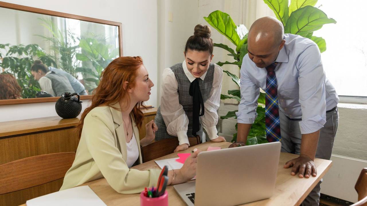 Three colleagues lean over a wooden office desk around a laptop and sticky notes during a working discussion.