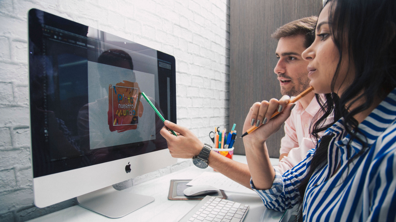 In-house creative team lead reviewing a project intake board and request queue on a monitor with designers at a shared desk in a modern brand office