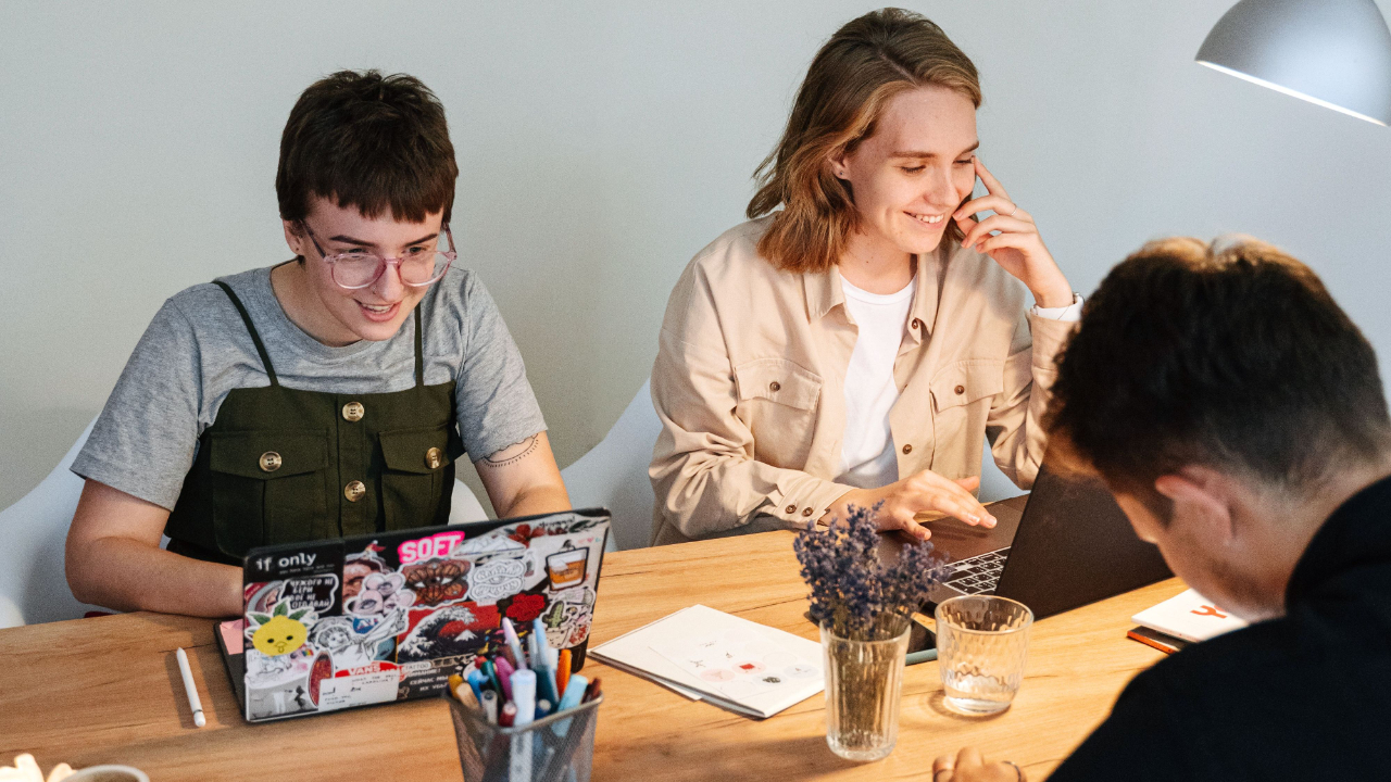 Design studio team collaborating on brand identity work and project plans at a shared desk with laptops, sketches, and design materials in a bright modern design studio
