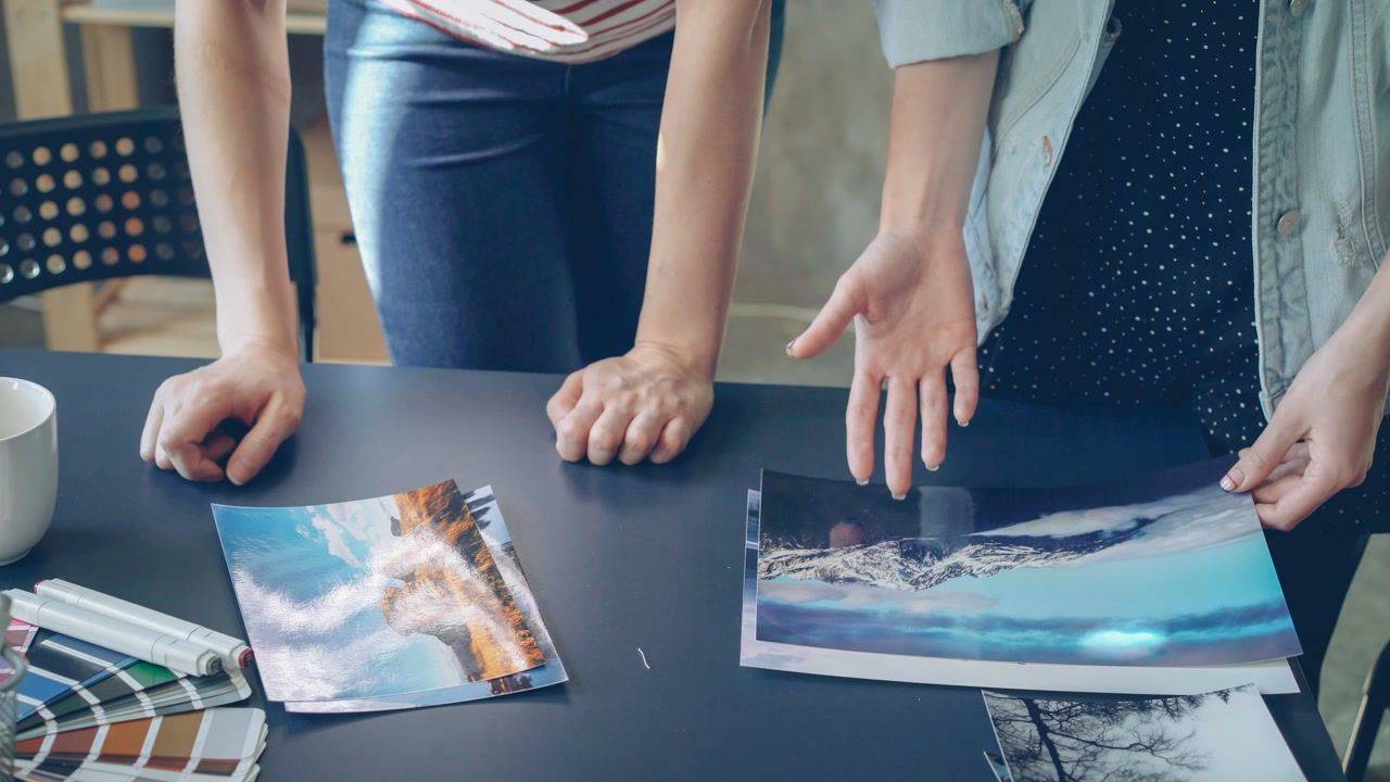 Two people stand over a dark studio table with printed landscape photographs, markers and a color swatch fan, hands gesturing at the prints during a review.