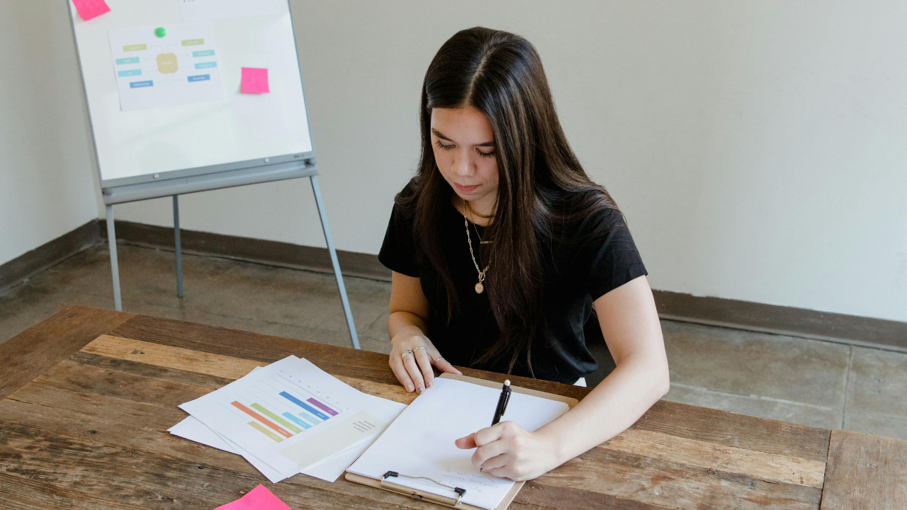Creative project manager writing notes and reviewing a brief document at a desk with laptop and printed materials in a bright modern agency office