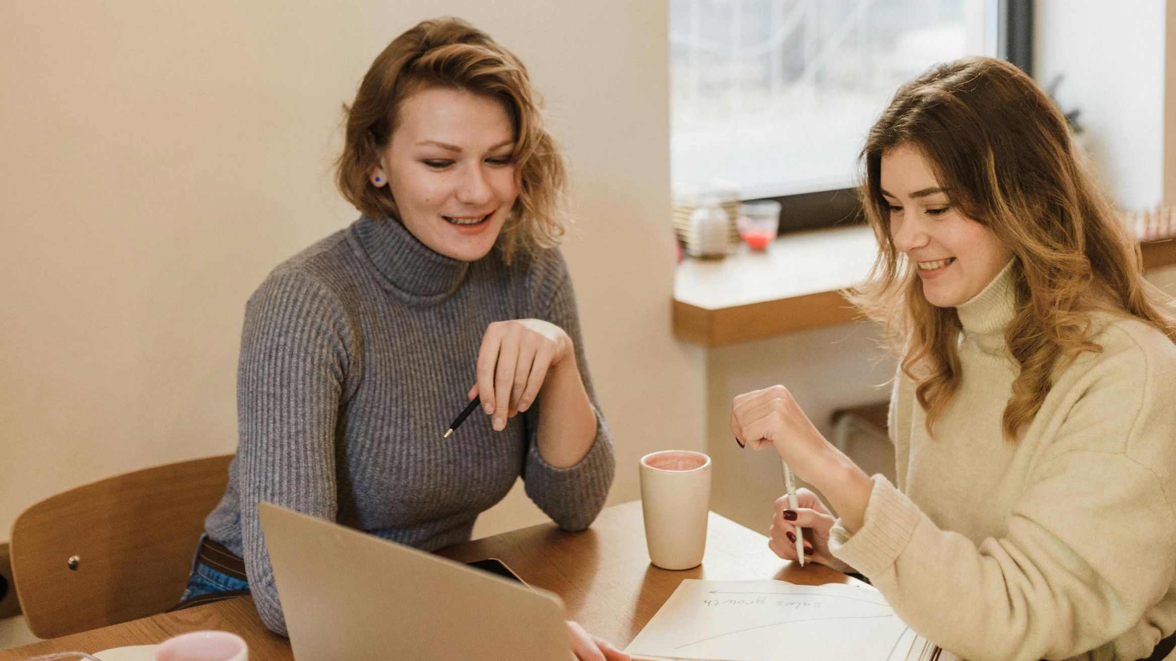 Two creative professionals comparing certification options on laptops at a modern design studio desk with notebooks and coffee