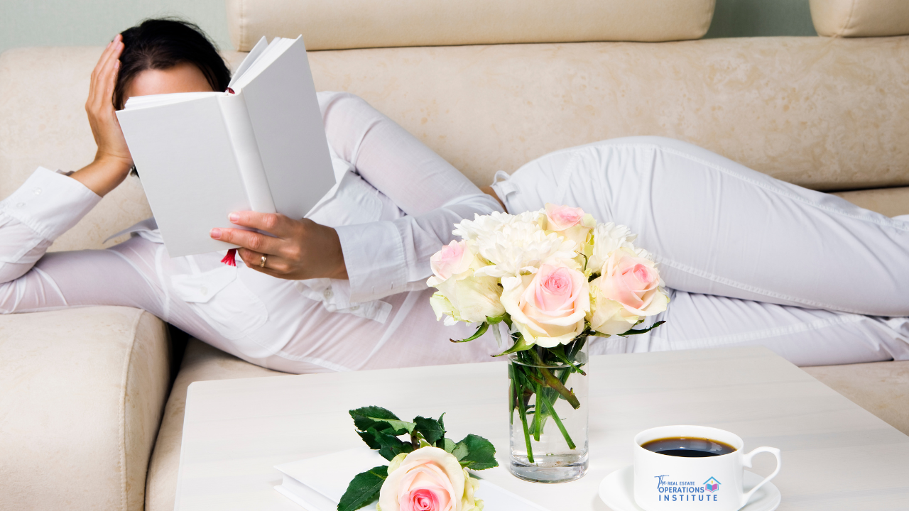 oman relaxing on couch reading a book with fresh flowers and coffee, representing rest and self-care for real estate professionals