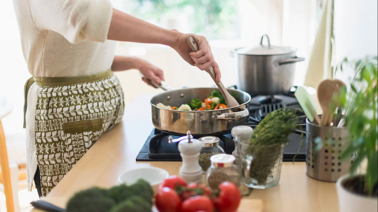 Person cooking vegetables in a pan on a stove. Kitchen setting with herbs, tomatoes, and utensils nearby. Bright, natural lighting.