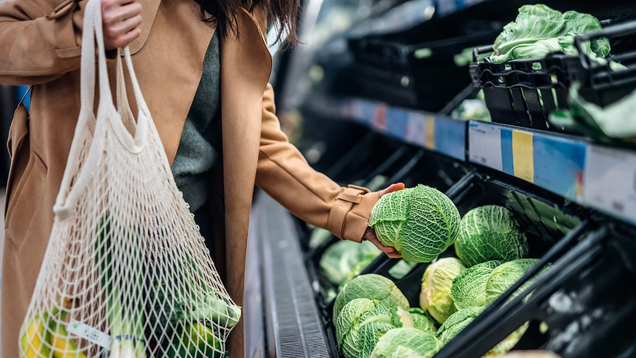 Person in a tan coat shops for cabbages at a grocery store, holding a mesh bag with bananas. Green cabbages line the shelves.