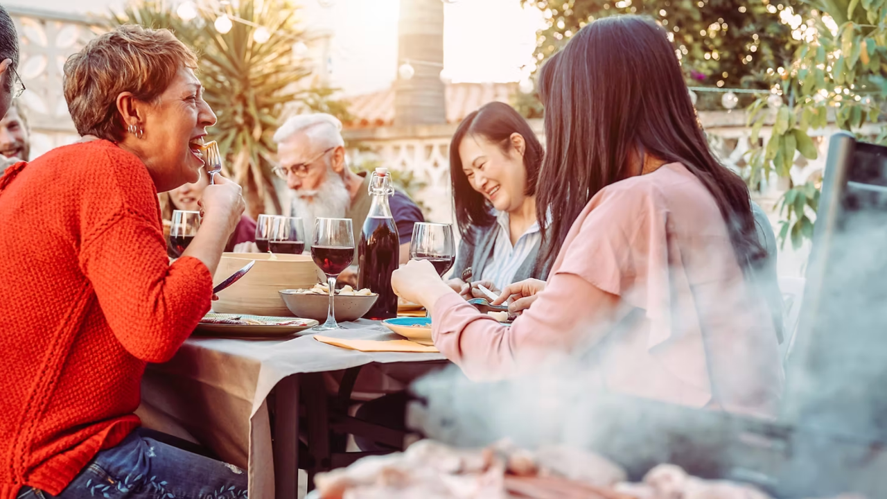 Group enjoying an outdoor meal, laughing around a table with wine glasses and food. BBQ smoke in foreground, greenery in background. Cozy mood.