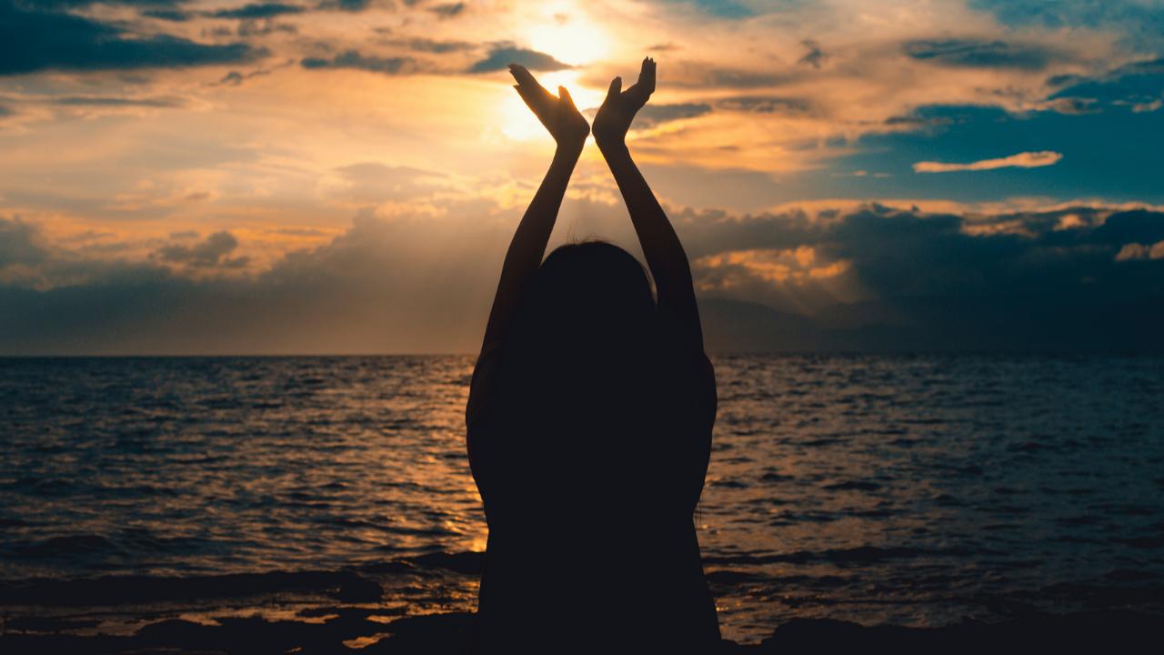 Silhouetted person raising their hands toward warm sunset light over the ocean, representing embodied language, physical cues, touch, and return in Cube Philosophy.