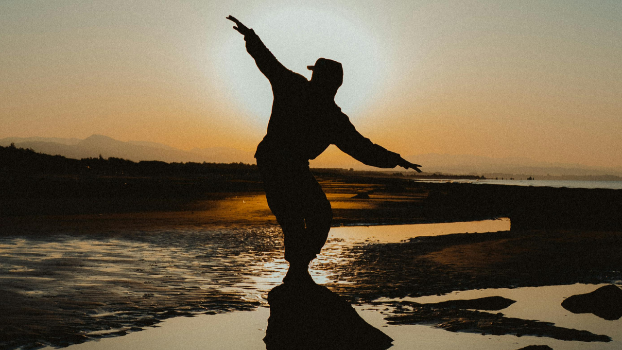 Silhouetted person balancing with outstretched arms beside reflective water at sunset, representing embodied orientation and the Cube becoming part of lived physical experience.