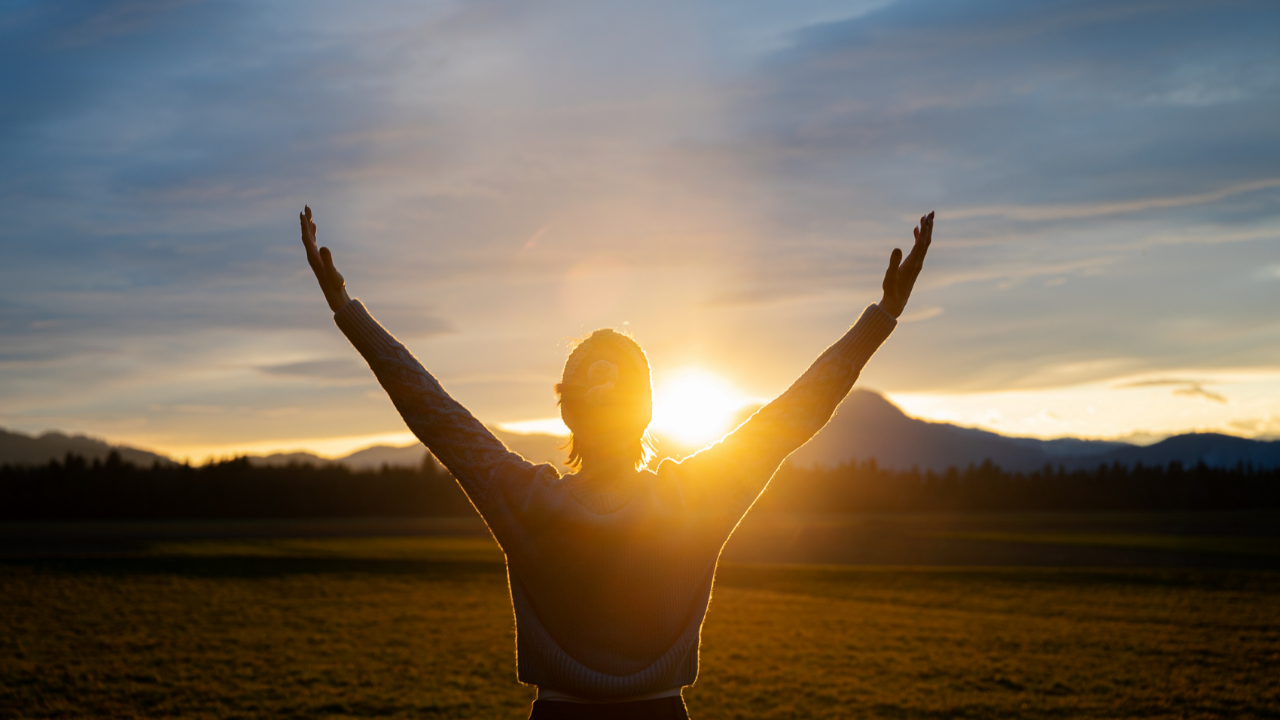 Silhouette of a woman standing with arms raised toward warm sunlight over a mountain landscape, representing release from bracing, embodied orientation, and return to Ground in Cube Philosophy.