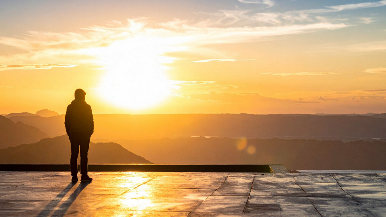 Silhouetted person standing before a brilliant golden sunrise over mountains, representing embodied orientation, practiced return, and learning the way back Home through the body.