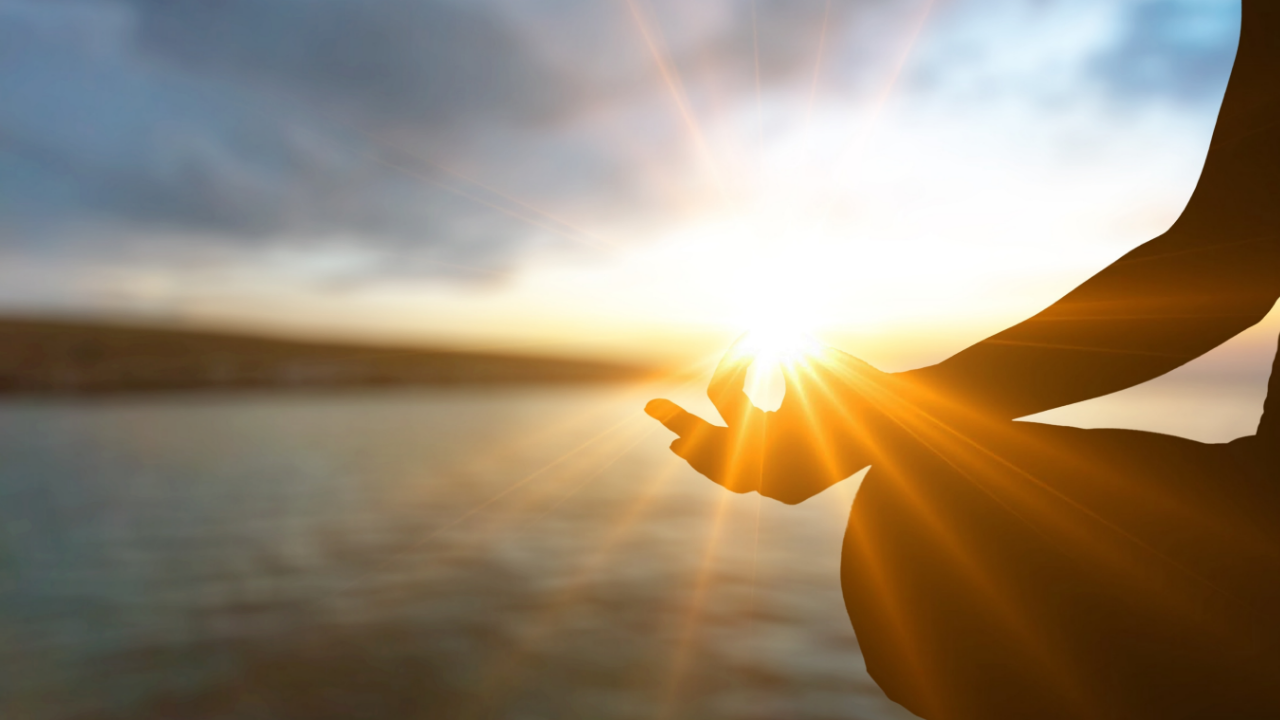 Silhouette of a person seated in stillness with hands open around bright sunlight, representing the pause between automatic reaction and conscious response in Cube Philosophy.